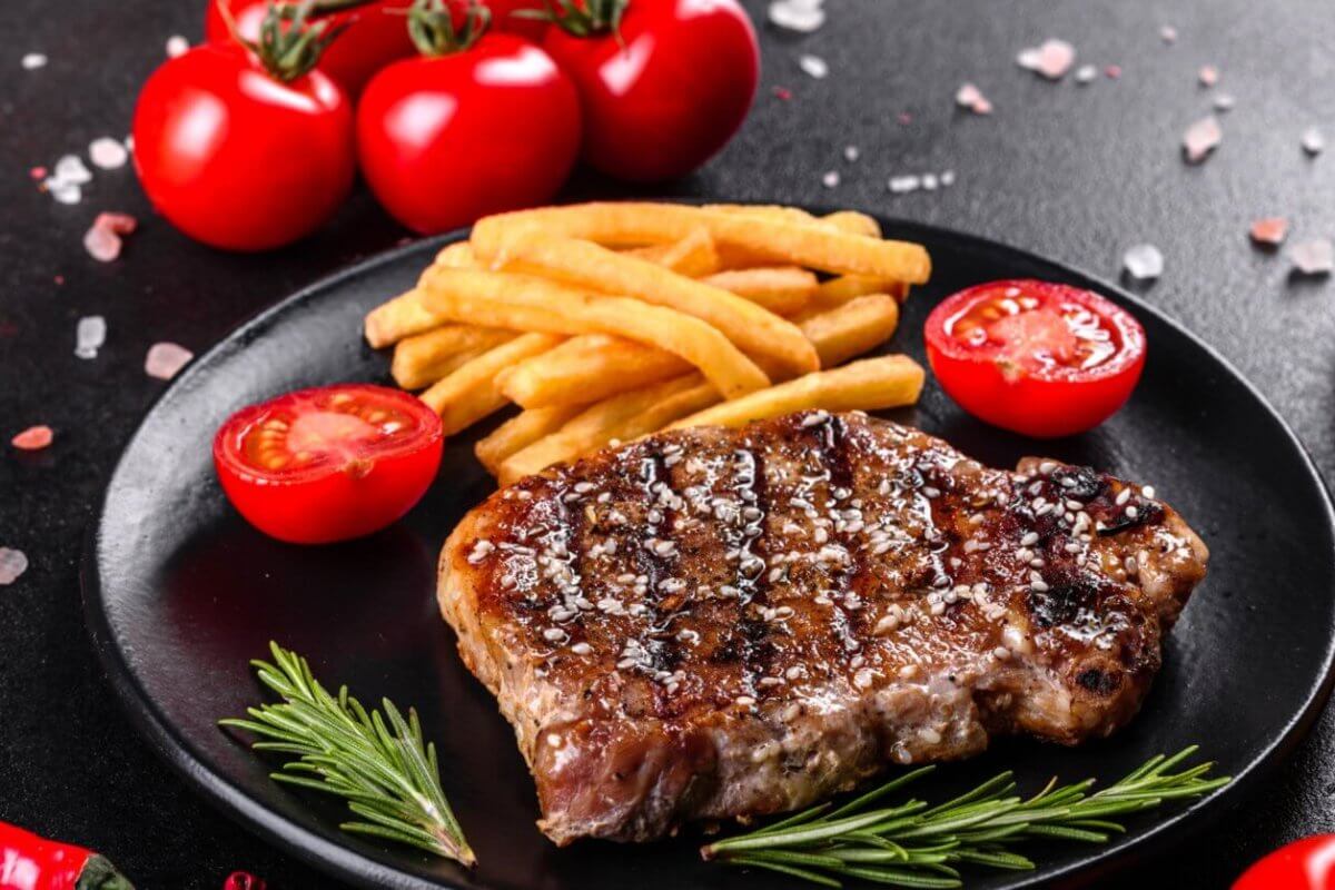 A grilled steak garnished with coarse salt and black pepper sits on a black plate alongside golden French fries and cherry tomato halves, promising a Sulit Dining Experience. Fresh rosemary sprigs lay next to the steak. The background features whole cherry tomatoes, a red chili pepper, sea salt, and a bottle of olive oil.