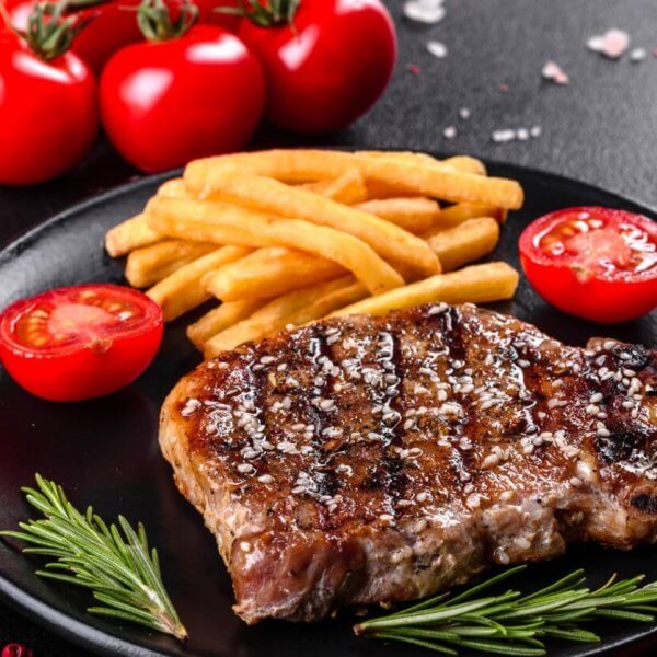 A grilled steak garnished with coarse salt and black pepper sits on a black plate alongside golden French fries and cherry tomato halves, promising a Sulit Dining Experience. Fresh rosemary sprigs lay next to the steak. The background features whole cherry tomatoes, a red chili pepper, sea salt, and a bottle of olive oil.