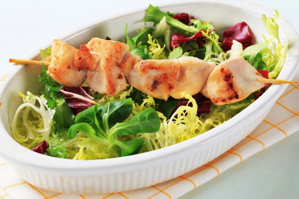 A white oval dish contains a fresh salad with green and purple lettuce, topped with a skewer of grilled chicken pieces. The dish, representing the best of sulit dining in Baguio, is placed on a folded, white napkin with yellow lines. The tender, slightly charred chicken contrasts with the vibrant greens of the salad.