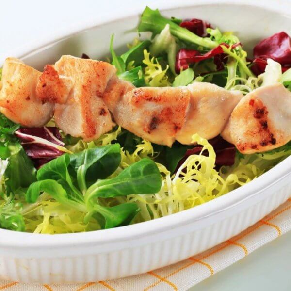 A white oval dish contains a fresh salad with green and purple lettuce, topped with a skewer of grilled chicken pieces. The dish, representing the best of sulit dining in Baguio, is placed on a folded, white napkin with yellow lines. The tender, slightly charred chicken contrasts with the vibrant greens of the salad.