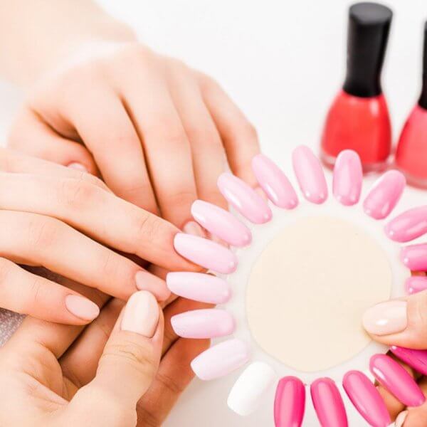 At one of the best nail lounges in Metro Manila, a person indulges in a relaxing manicure. Another holds a nail color palette of various pink and white shades against their nails. In the background, several bottles of red, pink, and beige nail polish sit on a pristine white surface, completing the spa-like ambiance.