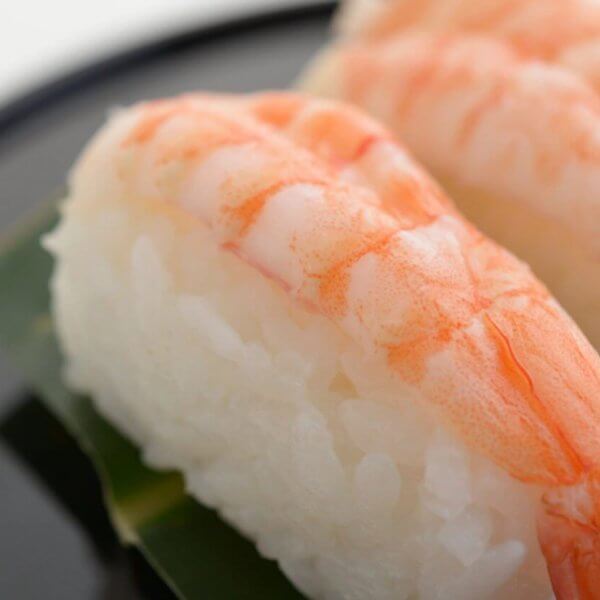 Close-up of two pieces of shrimp nigiri sushi on a black plate. Each sushi piece consists of a cooked shrimp placed atop a small mound of white rice, both resting on a green leaf. This tempting treat is part of the Food Lover's Guide at City of Dreams Manila. The background is blurred with a white backdrop.