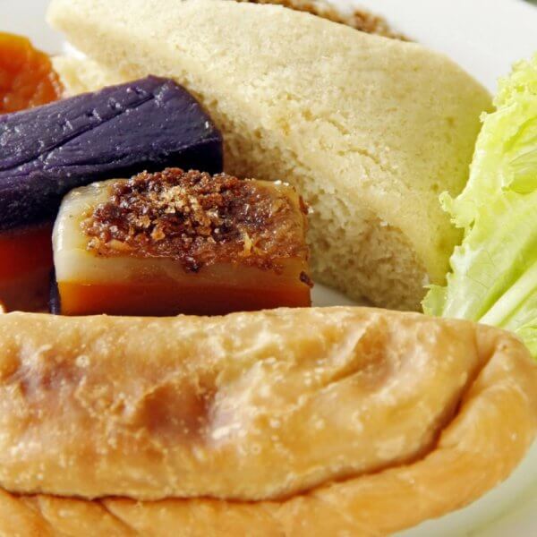 A plate with a variety of Filipino snacks, including a fried empanada in the foreground, slices of puto cake, sapin-sapin in purple and white, slices of orange kamote (sweet potato), and a piece of fresh lettuce on the side, all arranged on a white plate. Perfect for enjoying at one of Quezon City's best restaurants near Robinsons Magnolia.