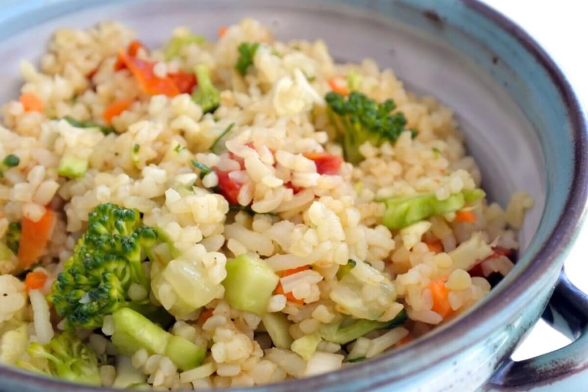 A ceramic bowl filled with a colorful vegetable rice dish, including broccoli florets, diced red bell peppers, and finely chopped carrots. The rice appears fluffy and well-cooked, with visible seasoning interspersed. Evoking the vibe of Katipunan's 63 Best Restaurants at UP Town Center, the bowl has rustic blue and gray tones.