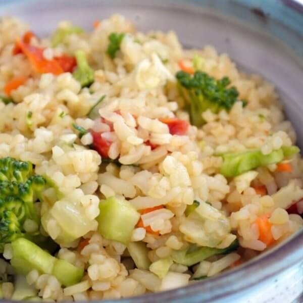 A ceramic bowl filled with a colorful vegetable rice dish, including broccoli florets, diced red bell peppers, and finely chopped carrots. The rice appears fluffy and well-cooked, with visible seasoning interspersed. Evoking the vibe of Katipunan's 63 Best Restaurants at UP Town Center, the bowl has rustic blue and gray tones.