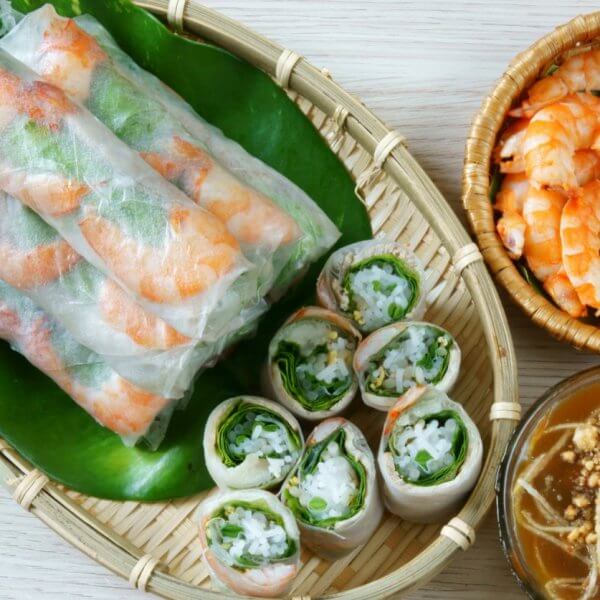 A woven tray holding fresh spring rolls filled with shrimp and vegetables rests atop a green banana leaf. Next to it, a small wicker bowl contains cooked shrimp, and another bowl is filled with brown peanut sauce garnished with crushed peanuts and sliced vegetables—a nod to authentic dining in Metro Manila's Vietnamese restaurants.