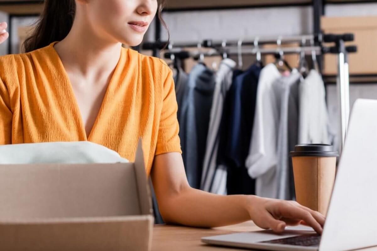 A woman in a yellow dress is sitting at a desk, holding a smartphone and typing on a laptop. In front of her is an open cardboard box, and a cup of coffee sits next to her computer. Behind her, a clothing rack with various garments is visible, suggesting she's running a small online business.