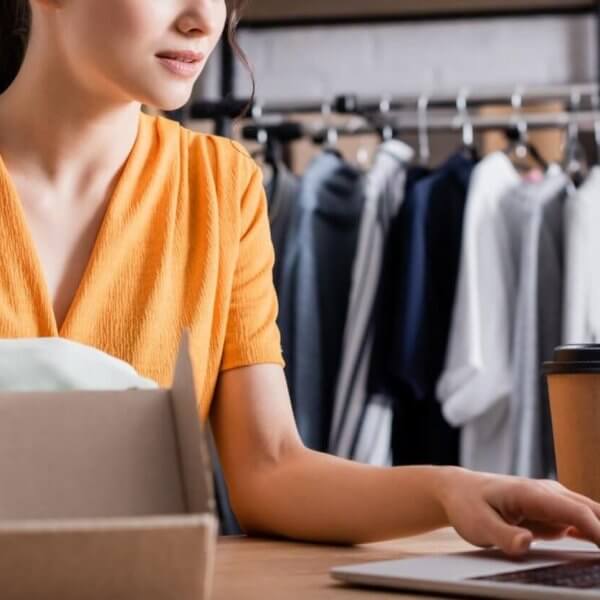 A woman in a yellow dress is sitting at a desk, holding a smartphone and typing on a laptop. In front of her is an open cardboard box, and a cup of coffee sits next to her computer. Behind her, a clothing rack with various garments is visible, suggesting she's running a small online business.
