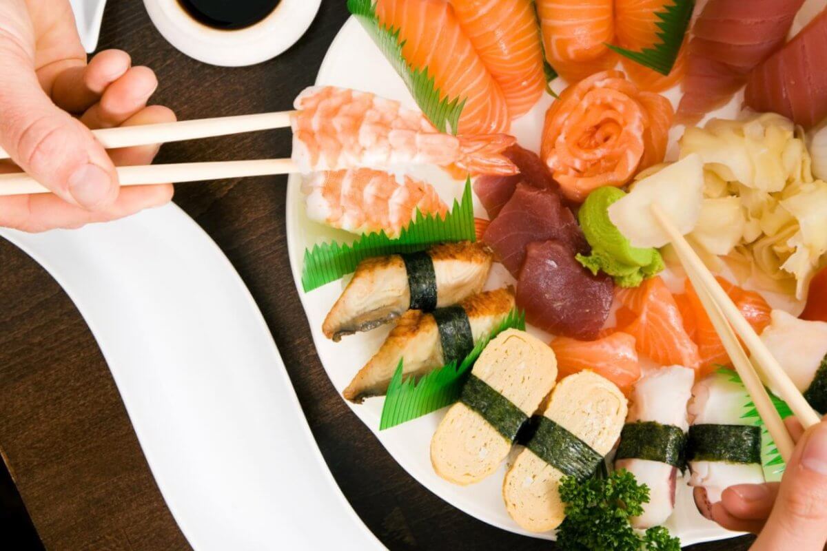 A variety of sushi and sashimi arranged on a white plate, including salmon, tuna, shrimp, and eel nigiri, tamago, and garnishes of wasabi, pickled ginger, and parsley. Two pairs of chopsticks are picking pieces from the platter at one of the restaurants in Ayala Alabang Town Center. A soy sauce dish is nearby on a dark wooden table.