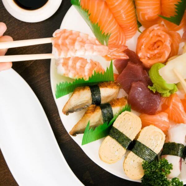 A variety of sushi and sashimi arranged on a white plate, including salmon, tuna, shrimp, and eel nigiri, tamago, and garnishes of wasabi, pickled ginger, and parsley. Two pairs of chopsticks are picking pieces from the platter at one of the restaurants in Ayala Alabang Town Center. A soy sauce dish is nearby on a dark wooden table.