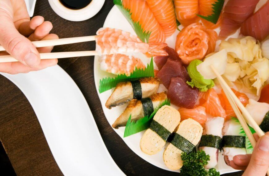 A variety of sushi and sashimi arranged on a white plate, including salmon, tuna, shrimp, and eel nigiri, tamago, and garnishes of wasabi, pickled ginger, and parsley. Two pairs of chopsticks are picking pieces from the platter at one of the restaurants in Ayala Alabang Town Center. A soy sauce dish is nearby on a dark wooden table.