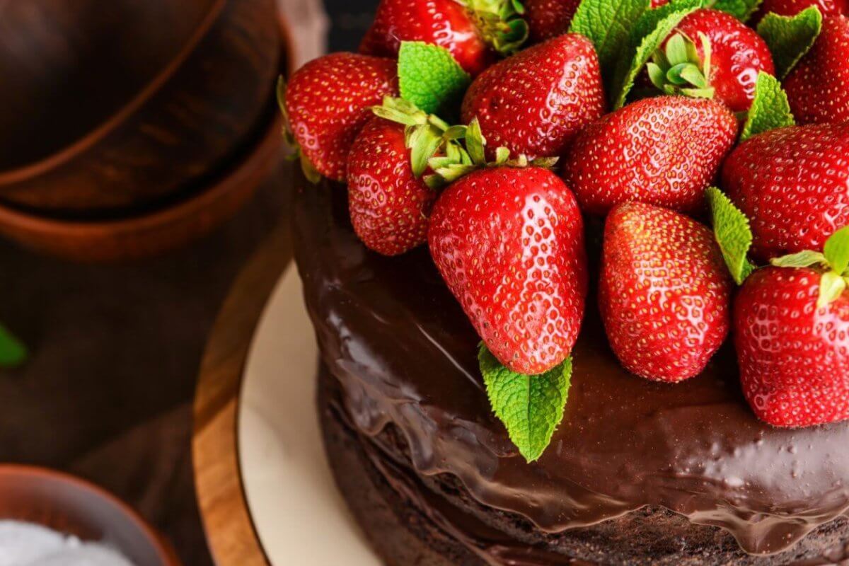 A close-up of a chocolate cake topped with a generous amount of fresh, red strawberries and green mint leaves. The cake has a shiny, smooth chocolate glaze. Brown bowls and green mint leaves are blurred in the background, perfect for those visiting the best restaurants at Ayala Feliz Mall.