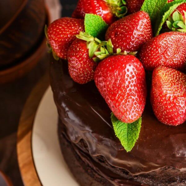 A close-up of a chocolate cake topped with a generous amount of fresh, red strawberries and green mint leaves. The cake has a shiny, smooth chocolate glaze. Brown bowls and green mint leaves are blurred in the background, perfect for those visiting the best restaurants at Ayala Feliz Mall.