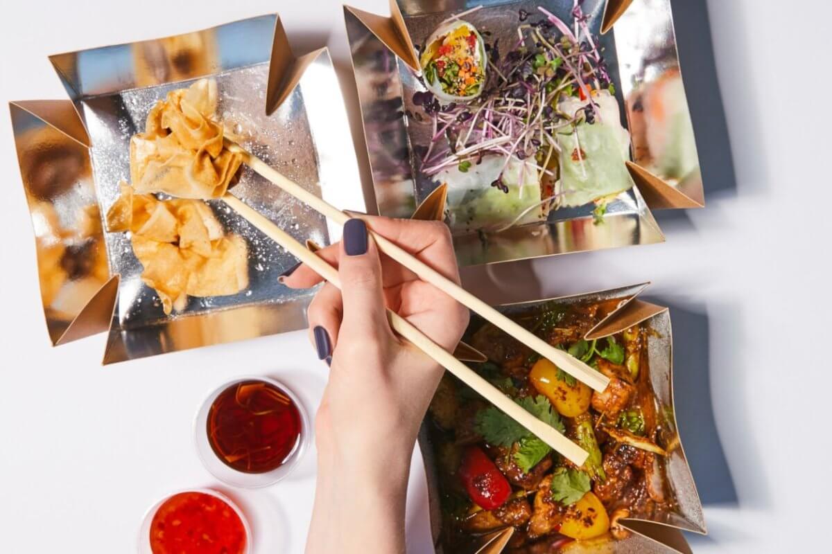 chopsticks to pick up a piece of food from one of three square, gold-colored dishes at Ayala Malls Vermosa. The dishes contain fried wontons, a salad with sprouts, and a cooked vegetable and meat dish. Two small dishes of red dipping sauces are placed nearby on a white surface.