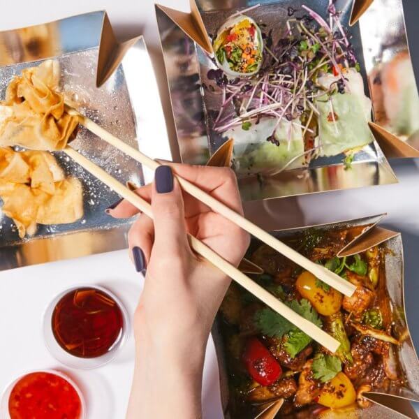 chopsticks to pick up a piece of food from one of three square, gold-colored dishes at Ayala Malls Vermosa. The dishes contain fried wontons, a salad with sprouts, and a cooked vegetable and meat dish. Two small dishes of red dipping sauces are placed nearby on a white surface.