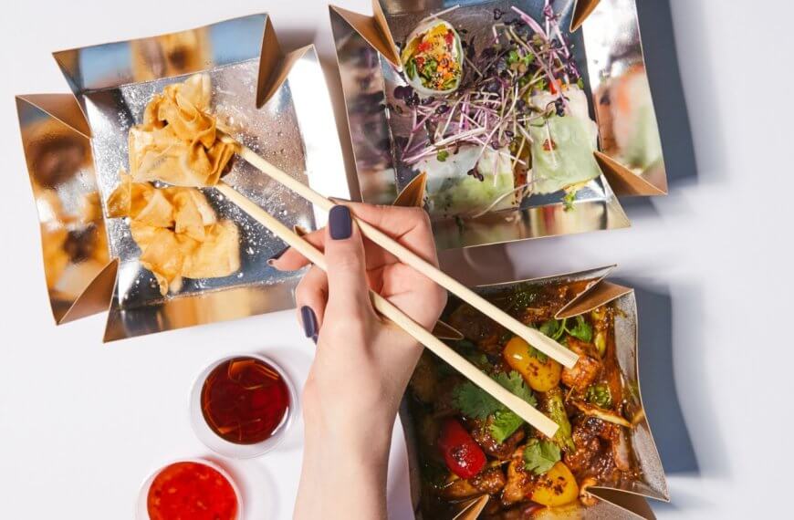 chopsticks to pick up a piece of food from one of three square, gold-colored dishes at Ayala Malls Vermosa. The dishes contain fried wontons, a salad with sprouts, and a cooked vegetable and meat dish. Two small dishes of red dipping sauces are placed nearby on a white surface.