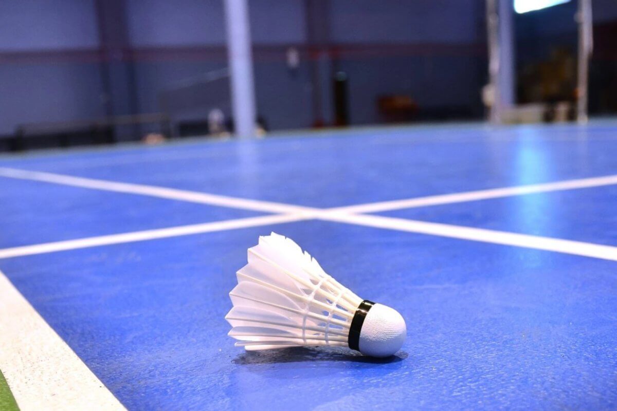 Close-up of a badminton shuttlecock lying on a blue indoor badminton court in Metro Manila. The shuttlecock, with its white feathers and black band, is in the foreground on the court's surface, with the net and other court lines visible in the background. The room has a blurred, spacious interior.