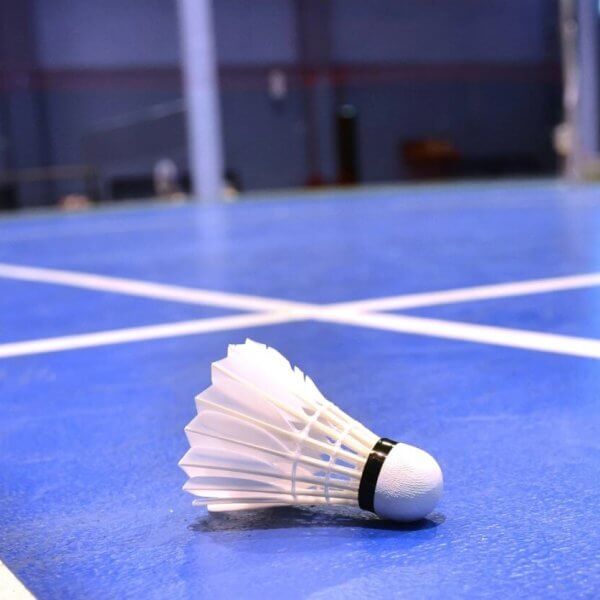 Close-up of a badminton shuttlecock lying on a blue indoor badminton court in Metro Manila. The shuttlecock, with its white feathers and black band, is in the foreground on the court's surface, with the net and other court lines visible in the background. The room has a blurred, spacious interior.