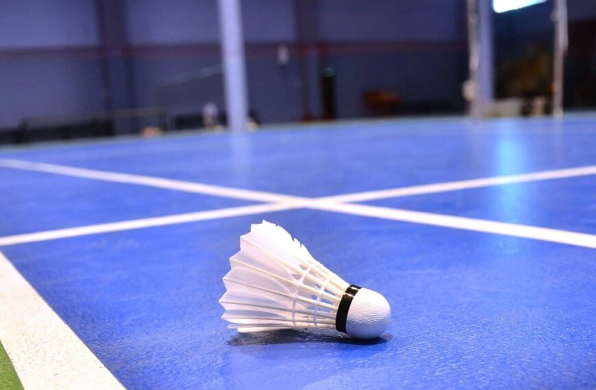 Close-up of a badminton shuttlecock lying on a blue indoor badminton court in Metro Manila. The shuttlecock, with its white feathers and black band, is in the foreground on the court's surface, with the net and other court lines visible in the background. The room has a blurred, spacious interior.