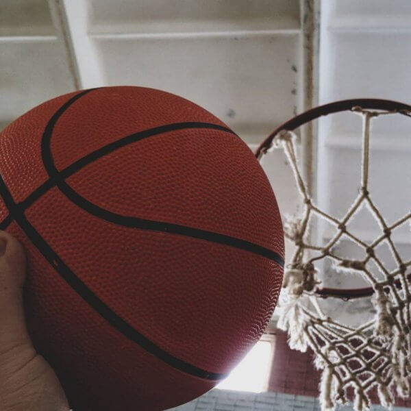 A hand holding an orange basketball is positioned near a hoop on an indoor basketball court available for rent.