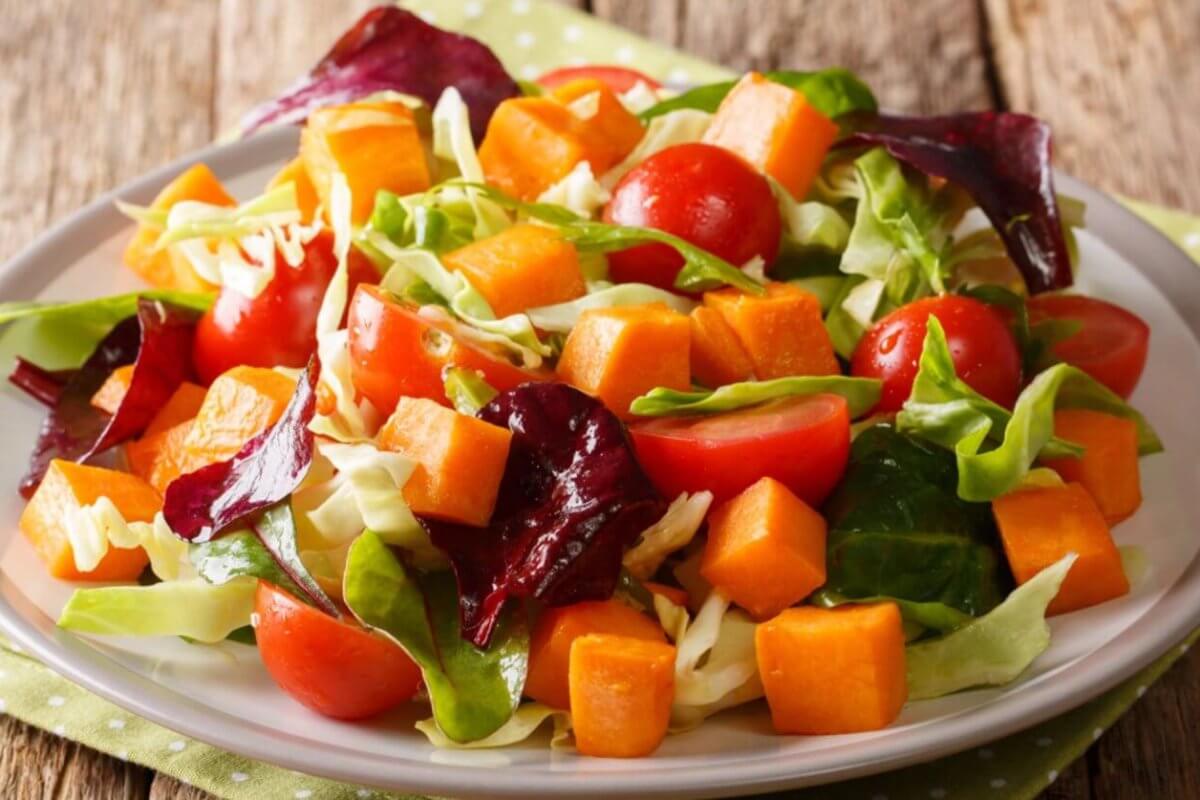 A fresh salad on a white plate features vibrant ingredients, including cherry tomatoes, cubed sweet potatoes, chopped cabbage, and a mix of green and purple leafy greens. Perfect for any food lover, this dish reflects the rich produce of Benguet Province. The plate rests on a green polka-dotted napkin on a rustic wooden table.