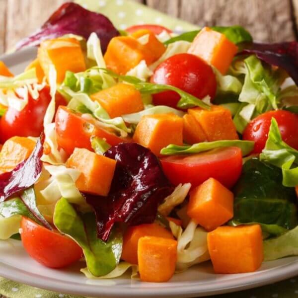A fresh salad on a white plate features vibrant ingredients, including cherry tomatoes, cubed sweet potatoes, chopped cabbage, and a mix of green and purple leafy greens. Perfect for any food lover, this dish reflects the rich produce of Benguet Province. The plate rests on a green polka-dotted napkin on a rustic wooden table.