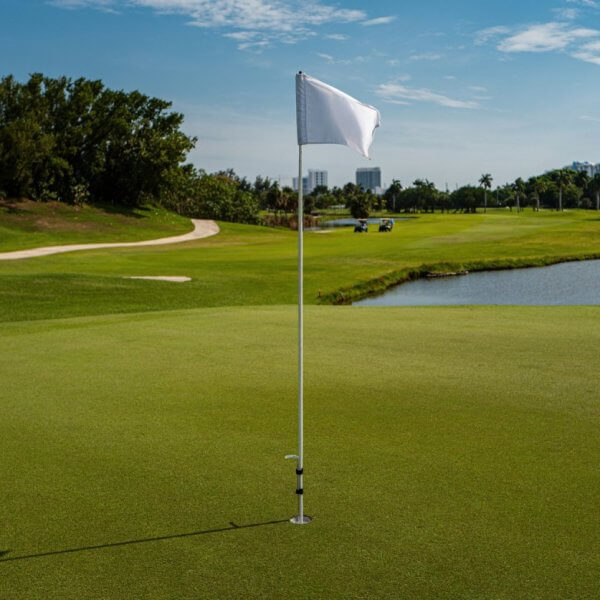 A golf course with a close-up view of a hole marked by a white flag on a green at one of the best golf and country clubs in the Philippines. In the background, there's a fairway surrounded by trees, a sand bunker, and a water area on the right. The sky is mostly clear with distant buildings visible beyond the trees.