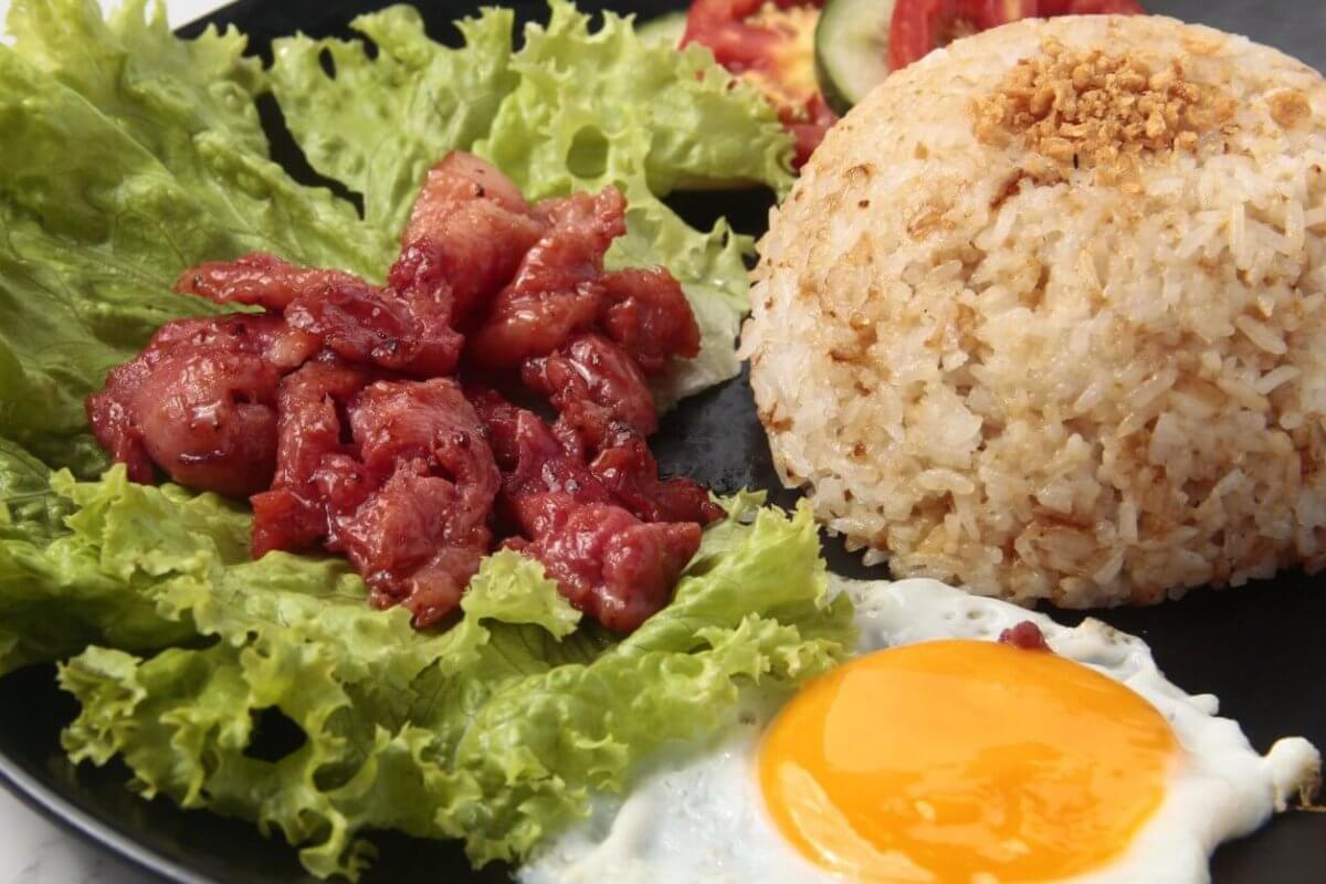 A black plate holds garlic fried rice topped with crushed garlic, accompanied by a sunny-side-up egg and pieces of caramelized pork placed over fresh green lettuce leaves. This classic Tapsilog, a popular Filipino breakfast in Metro Manila, is garnished with slices of tomato and cucumber in the background.