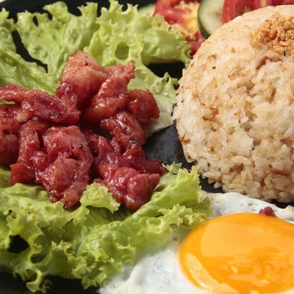 A black plate holds garlic fried rice topped with crushed garlic, accompanied by a sunny-side-up egg and pieces of caramelized pork placed over fresh green lettuce leaves. This classic Tapsilog, a popular Filipino breakfast in Metro Manila, is garnished with slices of tomato and cucumber in the background.