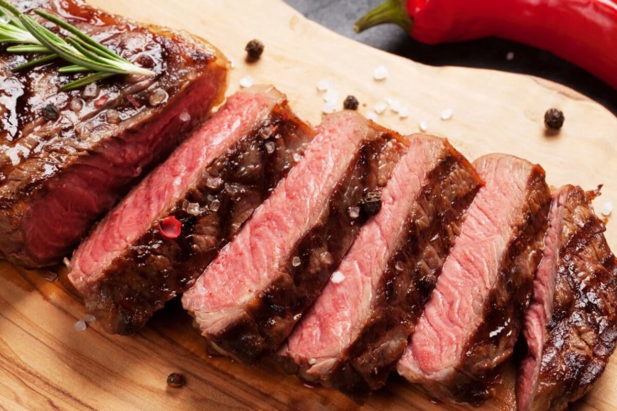 A cooked steak, partially sliced into pieces, is presented on a wooden cutting board, garnished with coarse sea salt and black peppercorns. A sprig of rosemary rests on the whole part of the steak. In the background at Bonifacio High Street's top restaurants in BGC, whole red and yellow chili peppers are visible.