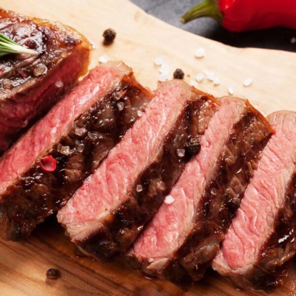 A cooked steak, partially sliced into pieces, is presented on a wooden cutting board, garnished with coarse sea salt and black peppercorns. A sprig of rosemary rests on the whole part of the steak. In the background at Bonifacio High Street's top restaurants in BGC, whole red and yellow chili peppers are visible.