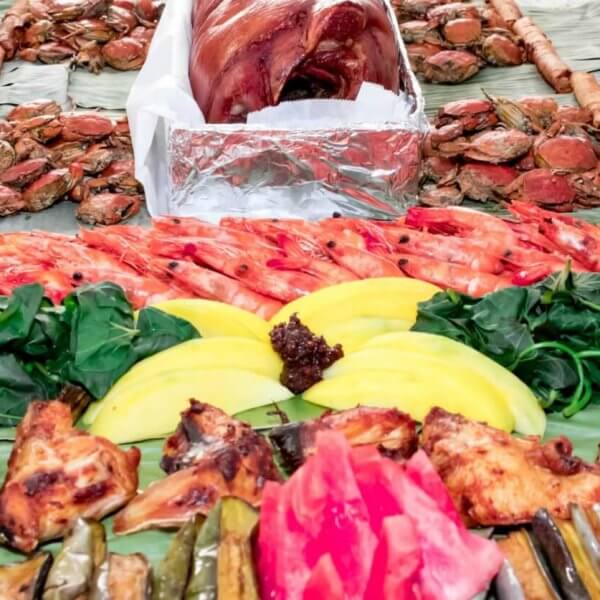 A festive spread of Filipino food is served on banana leaves at one of Metro Manila's top Boodle Fight Restaurants. Central to the layout are roasted pork, grilled chicken, shrimp, and vibrant fruits like mango and watermelon. Accompanying these are fresh greens, crabs, various grilled vegetables, and spring rolls arranged in an inviting manner.