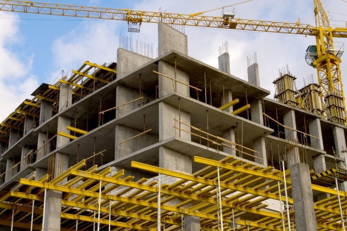 A construction site features a multi-story concrete building framework. Prominently visible are yellow scaffolding and support structures. Two tall tower cranes, essential for construction companies in the Philippines, are stationed nearby, one crane's arm extending over the building. The sky is partly cloudy with blue patches.