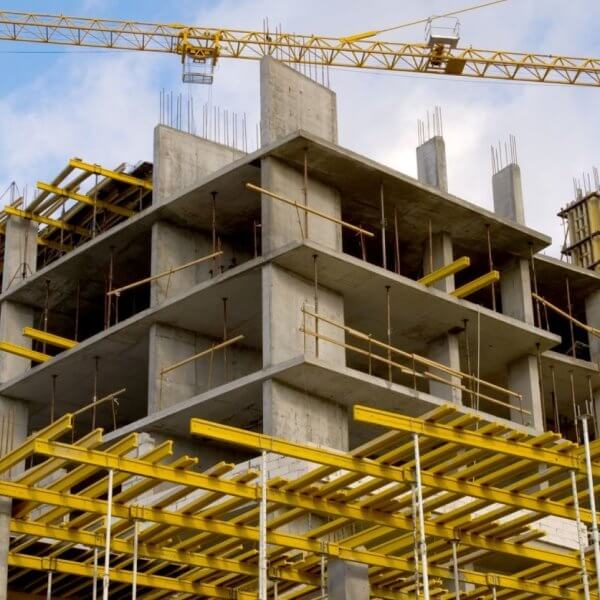 A construction site features a multi-story concrete building framework. Prominently visible are yellow scaffolding and support structures. Two tall tower cranes, essential for construction companies in the Philippines, are stationed nearby, one crane's arm extending over the building. The sky is partly cloudy with blue patches.
