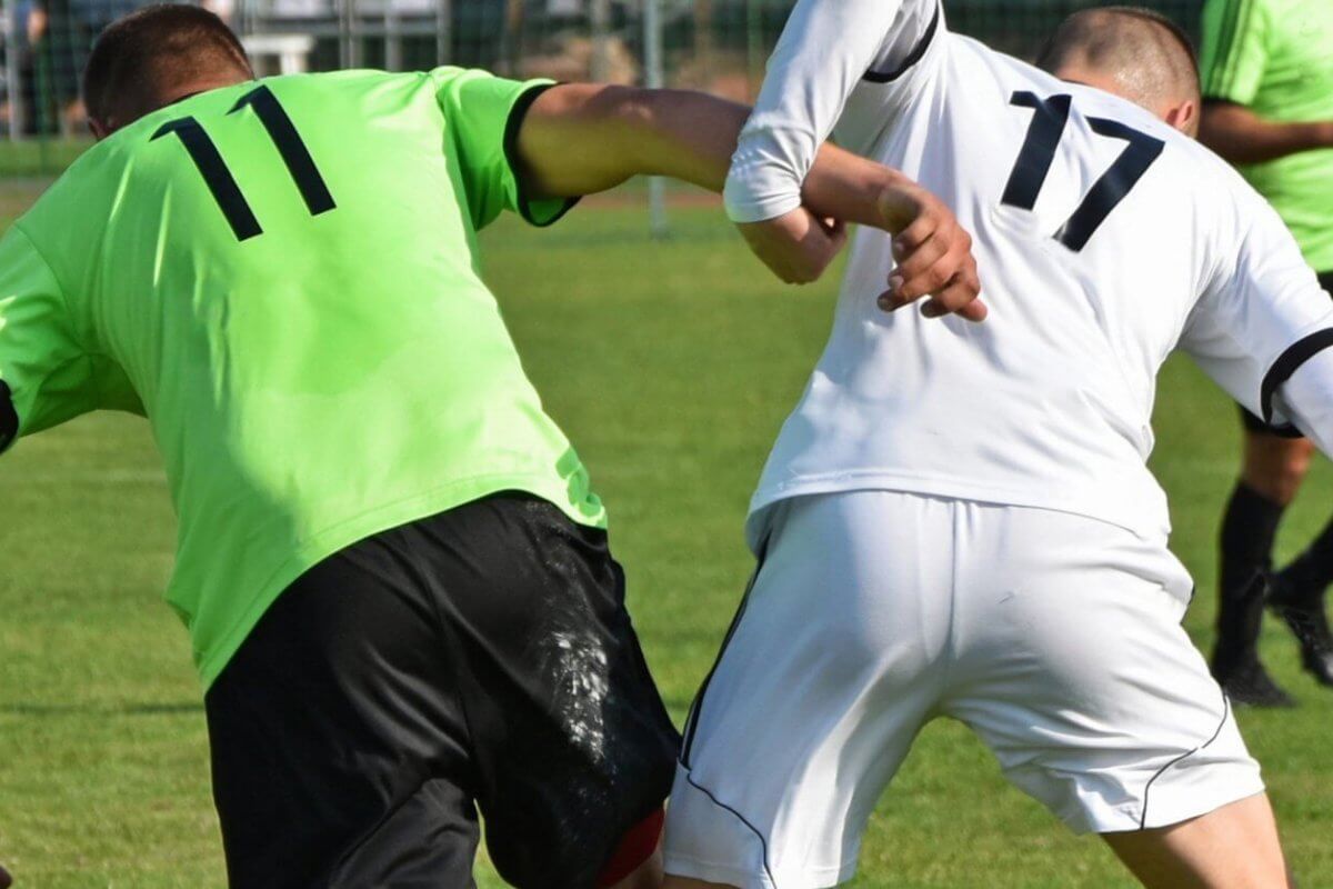 Two soccer players, each showcasing custom uniforms—one in a green jersey with the number 11 and the other in a white jersey with the number 17—compete for the ball on a grassy field. Both are leaning in and making contact with their arms as they balance on one leg, underlining their suppliers' quality craftsmanship. A goal post and fencing are visible in the background.