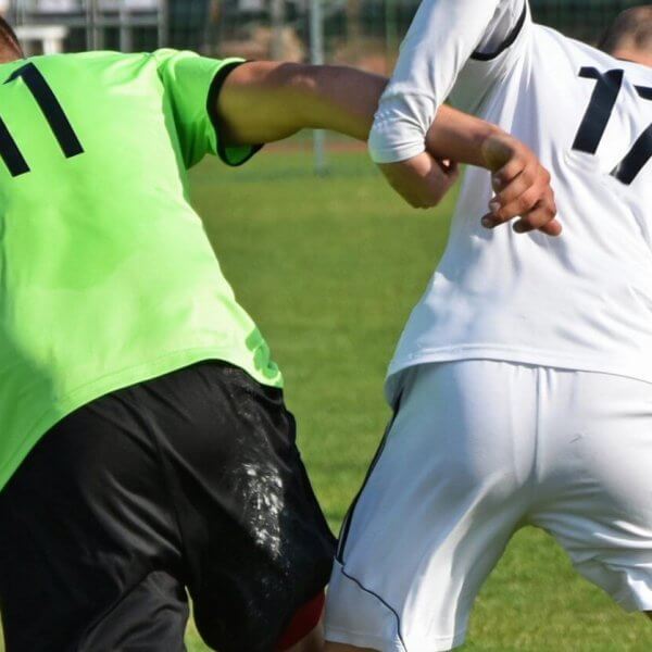 Two soccer players, each showcasing custom uniforms—one in a green jersey with the number 11 and the other in a white jersey with the number 17—compete for the ball on a grassy field. Both are leaning in and making contact with their arms as they balance on one leg, underlining their suppliers' quality craftsmanship. A goal post and fencing are visible in the background.