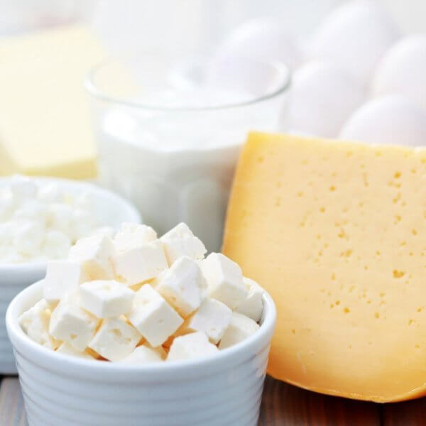 A wooden table in Metro Manila showcases a variety of dairy products supplied by local Dairy Product Suppliers, including a block of cheddar cheese, a bowl of white feta cheese cubes, cottage cheese, glass of milk, dish of butter, and a bowl of eggs. The items are elegantly arranged in white bowls and on plates.