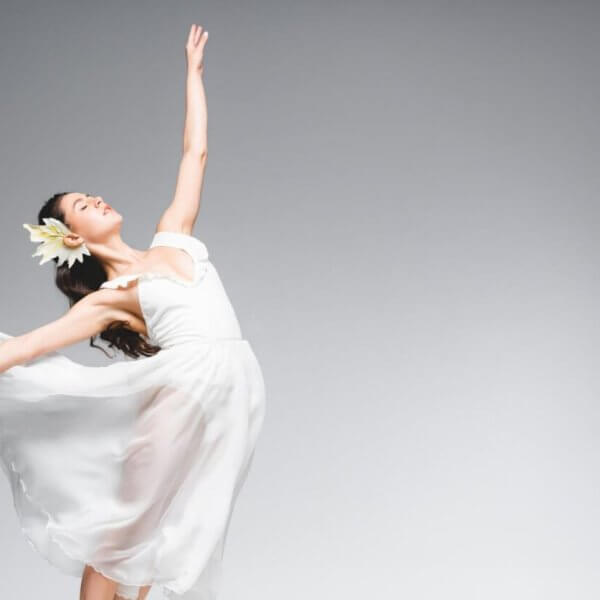 A ballet dancer in a flowing white dress and flower headpiece gracefully poses against a plain grey background. Her dress billows around her as she stands en pointe, creating a sense of motion and elegance—a true representation of the best dance studios in Metro Manila.