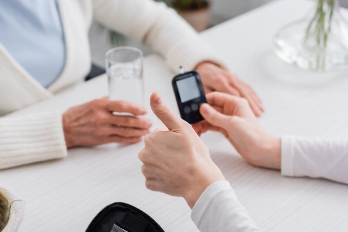 Two people sit at a white table in a bustling Metro Manila clinic. One person holds a blood glucose meter, giving a thumbs-up, while the other holds a glass of water. On the table are a zippered case with medical supplies and a bowl with green apples. The background is blurred, focusing on their interaction.