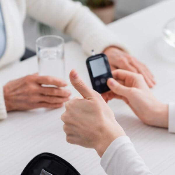 Two people sit at a white table in a bustling Metro Manila clinic. One person holds a blood glucose meter, giving a thumbs-up, while the other holds a glass of water. On the table are a zippered case with medical supplies and a bowl with green apples. The background is blurred, focusing on their interaction.