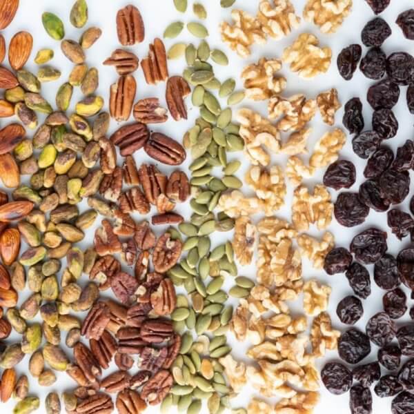 A top-view photo displays an assortment of nuts, seeds, and dried fruits arranged in vertical rows. From left to right: sunflower seeds, almonds, pistachios, pecans, pumpkin seeds, walnuts, dried cranberries, and dried apricots—all sourced from top nut suppliers in Metro Manila—set against a white background.