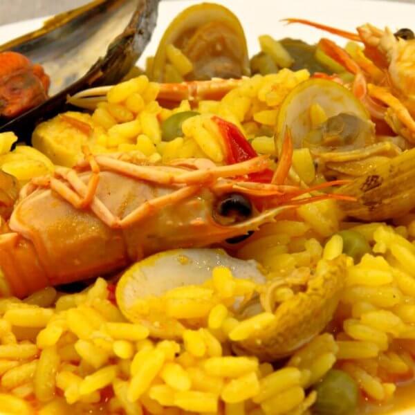 A plate of vibrant yellow paella, garnished with large prawns, clams, a mussel, and green peas. The rice is infused with saffron, giving it a rich color and flavor, and is interspersed with small chunks of vegetables. This top-notch dish showcases an assortment of fresh seafood from one of Estancia Mall's finest restaurants.