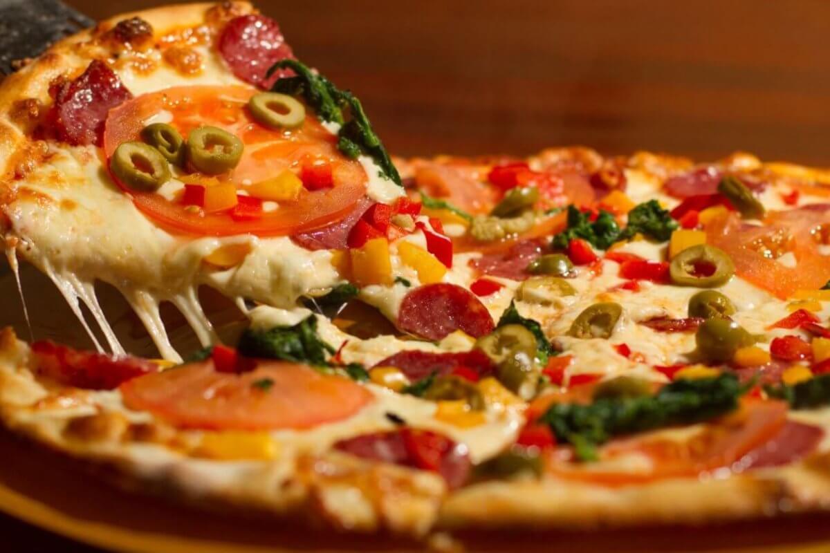 pizza at one of the best restaurants in Festival Mall. The pizza is topped with melted cheese, pepperoni, tomato slices, green olives, red bell peppers, and leafy greens. The crust is golden brown, and the cheese is stretchy as the slice is pulled away.