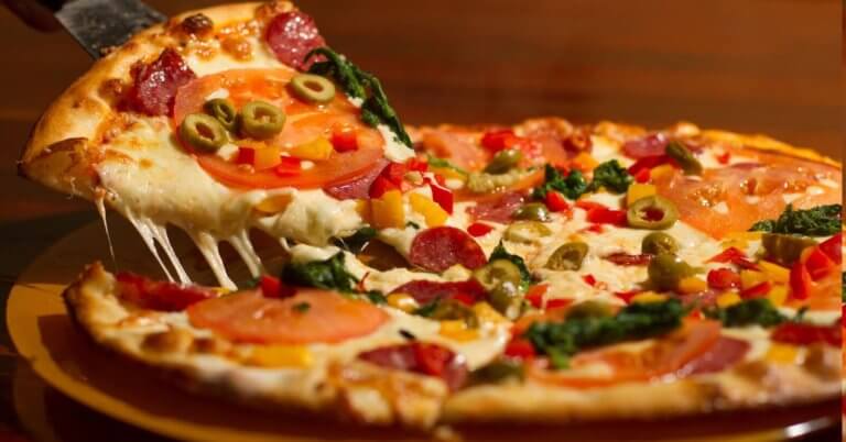 pizza at one of the best restaurants in Festival Mall. The pizza is topped with melted cheese, pepperoni, tomato slices, green olives, red bell peppers, and leafy greens. The crust is golden brown, and the cheese is stretchy as the slice is pulled away.