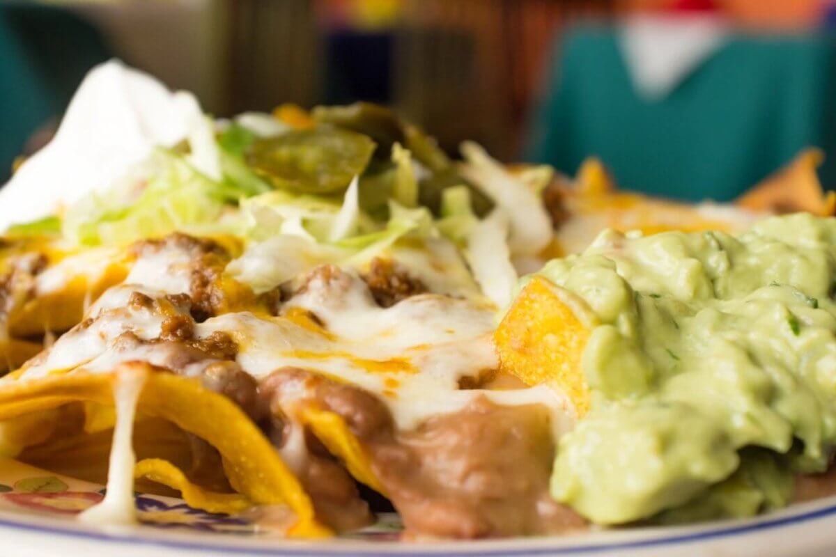 nachos topped with melted cheese, refried beans, ground beef, shredded lettuce, jalapeños, and a dollop of sour cream. On the side is a generous portion of creamy guacamole. The background is blurred to highlight the focus on these FIL-MEX delights from Metro Manila.
