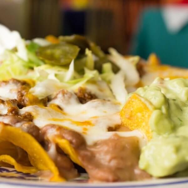 nachos topped with melted cheese, refried beans, ground beef, shredded lettuce, jalapeños, and a dollop of sour cream. On the side is a generous portion of creamy guacamole. The background is blurred to highlight the focus on these FIL-MEX delights from Metro Manila.