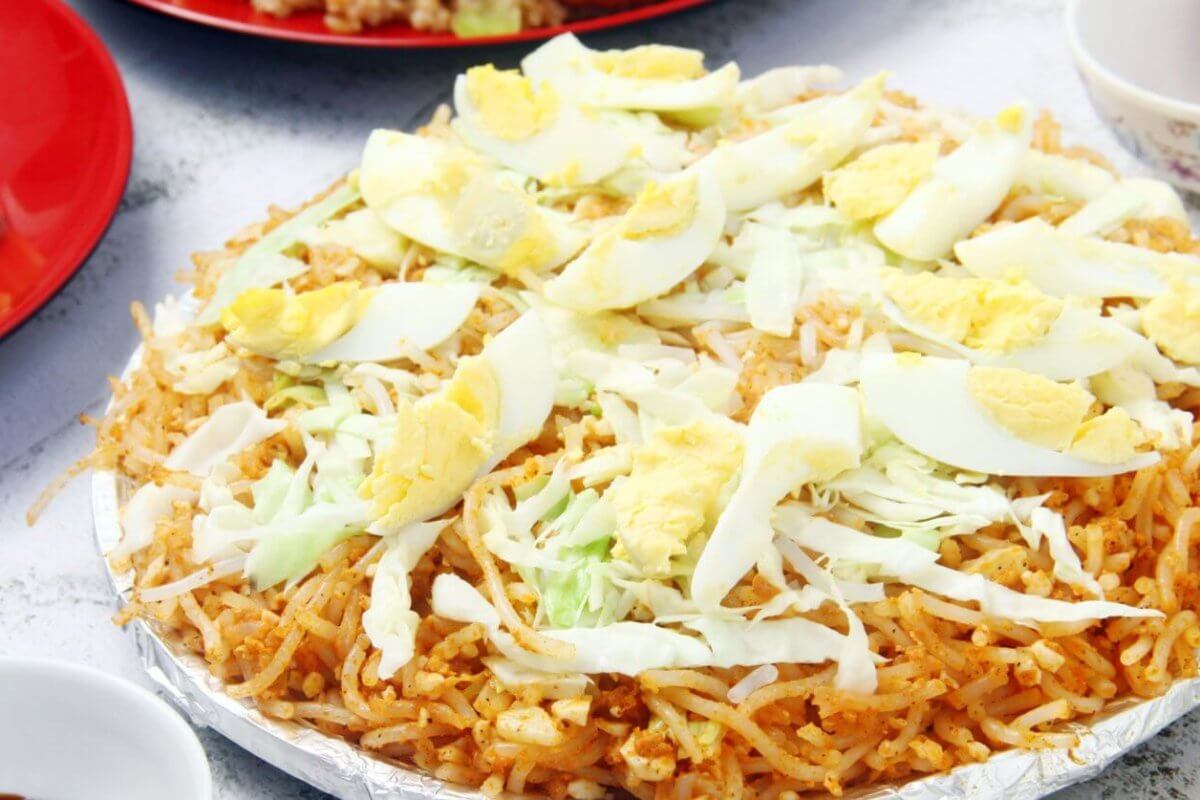A plate of Filipino pancit palabok is topped with sliced hard-boiled eggs, shredded cabbage, and bits of garlic. The yellow-orange noodles are seasoned and mixed with shrimp sauce. Nearby, there are partially visible plates and bowls with other dishes and condiments on a white textured surface, reminiscent of the vibrant food parks in Metro Manila.