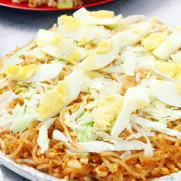 A plate of Filipino pancit palabok is topped with sliced hard-boiled eggs, shredded cabbage, and bits of garlic. The yellow-orange noodles are seasoned and mixed with shrimp sauce. Nearby, there are partially visible plates and bowls with other dishes and condiments on a white textured surface, reminiscent of the vibrant food parks in Metro Manila.