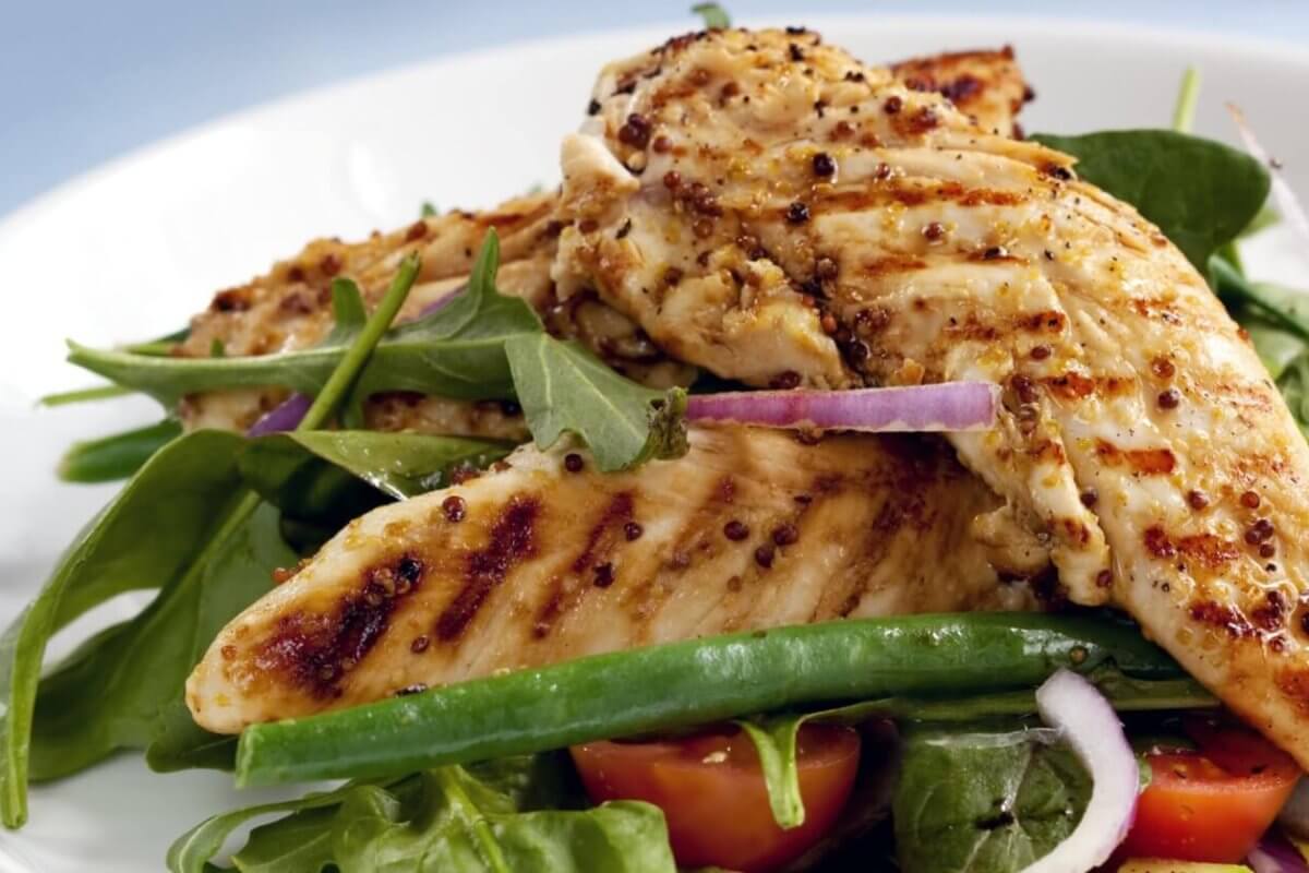 A plate of grilled chicken breasts served on a bed of fresh mixed greens, including arugula and green beans, is now available through healthy food delivery in Manila. The dish is garnished with slices of red onion and halves of cherry tomatoes. The chicken has a seared, seasoned exterior, and the salad appears crisp and vibrant.