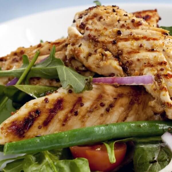 A plate of grilled chicken breasts served on a bed of fresh mixed greens, including arugula and green beans, is now available through healthy food delivery in Manila. The dish is garnished with slices of red onion and halves of cherry tomatoes. The chicken has a seared, seasoned exterior, and the salad appears crisp and vibrant.